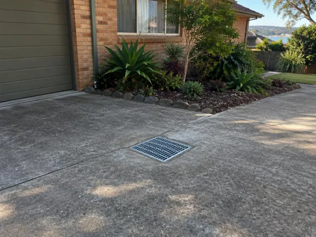 Concrete driveway with drain grate beside a brick house and garden bed in Narrabeen